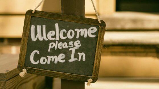 Sign that says "Welcome please come in." White script text on black background with wooden frame hangs by rope on a porch post